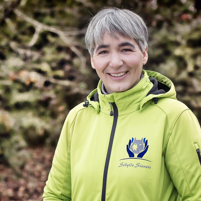 Sibylle Sievers Eine lächelnde Frau (Sibylle Sievers) in einer grünen Jacke steht vor einer natürlichen Kulisse.